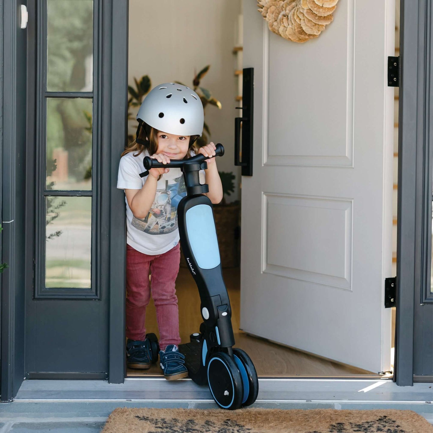 Child wearing helmet coming our of front door smiling pushing blue Larktale Scoobi in scooter mode