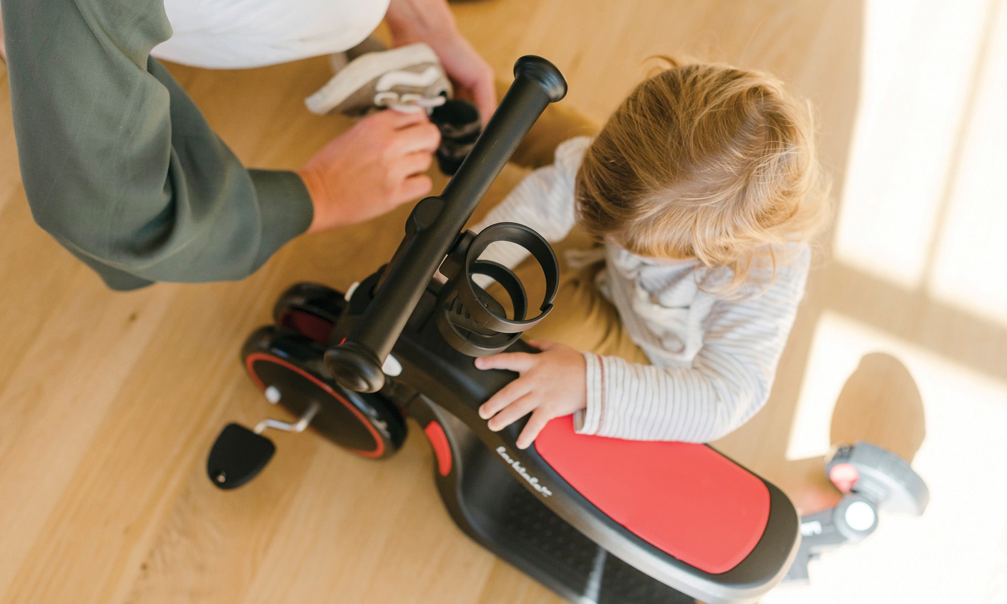 parent putting on toddlers shoes whilst baby is playing with red tricycle