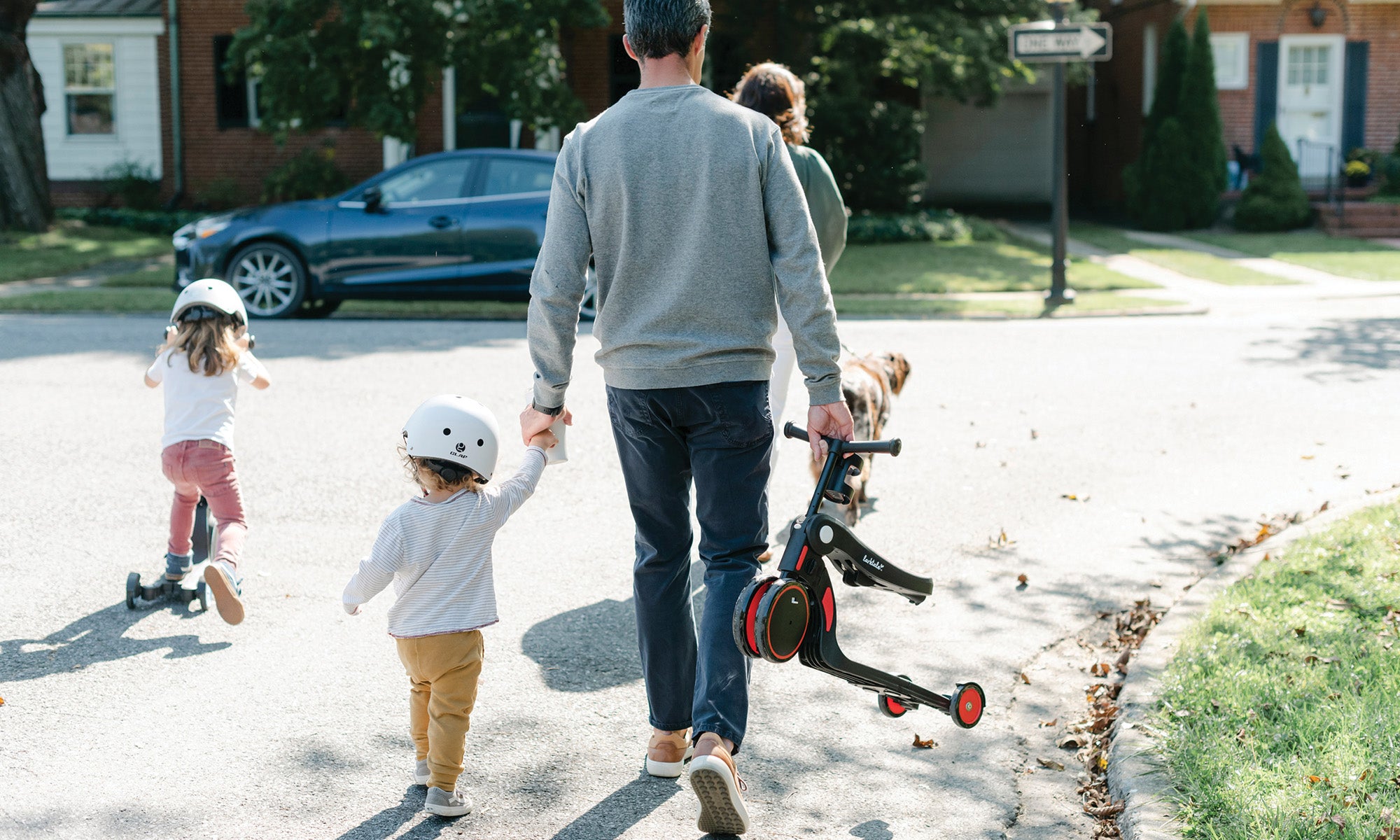 family walking dog with two toddlers playing on their scooters wearing helmets 
